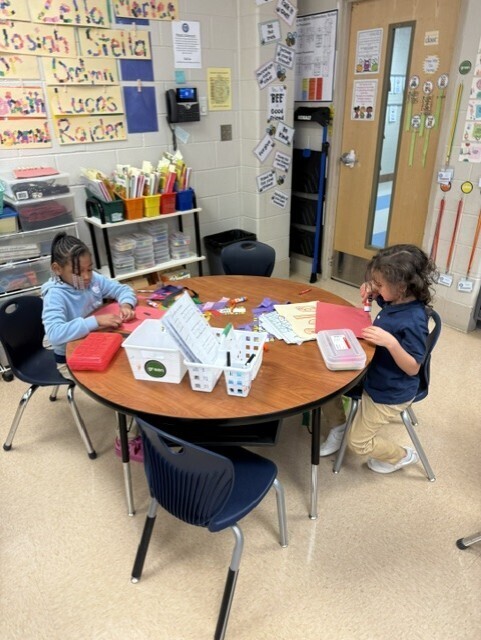 Two children seated around a small table in a classroom, working on crafts. Brightly colored papers and materials are on the table.