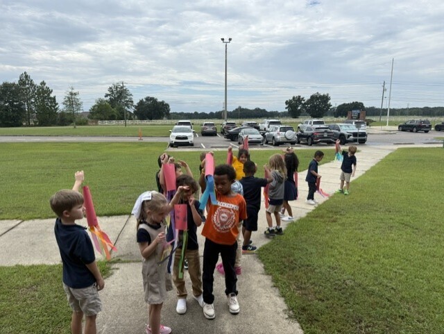 Children stand on a path with ribbons, a grassy field and parked cars in the background.