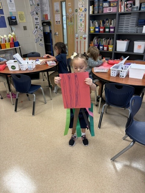 Child stands in classroom, holding red paper in both hands, surrounded by tables with supplies.