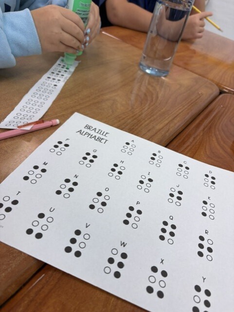 Child seated at a desk with a Braille worksheet, a glass of water, and a pencil.