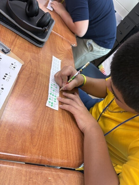 A student uses a pencil to complete a Braille worksheet at a desk, with headphones and another student in the background.