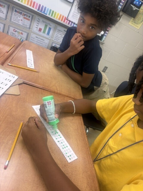 Two children in a classroom. One is applying glue to a paper strip, the other sits nearby.