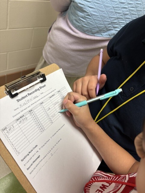 Child holds a clipboard with a pen, working on a recording sheet. They wear a lanyard and a red shirt.