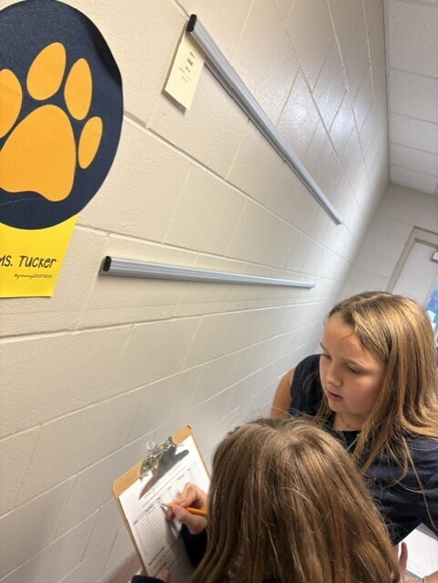Two students, one writing, in a hallway with a paw print and a sign reading "Ms. Tucker" on the wall.