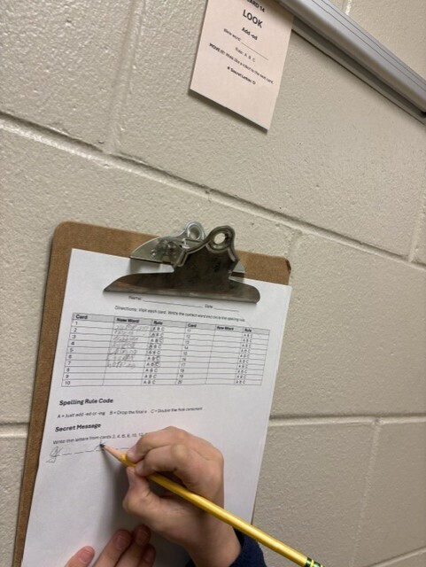 A person writes on a paper attached to a clipboard on a wall. A note reads "Look".