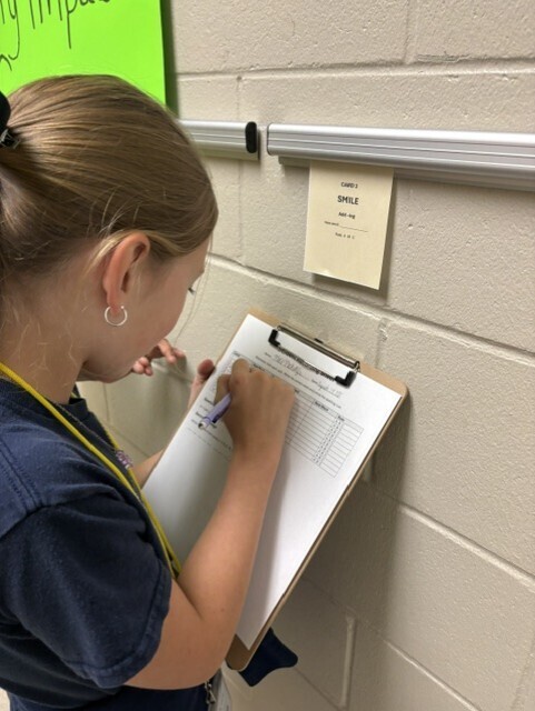 A person writing on a clipboard against a wall with a yellow sign attached to the wall.