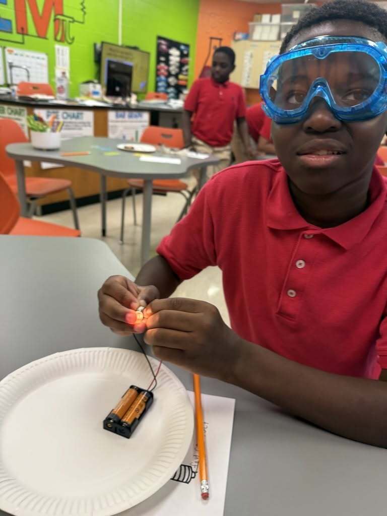 A boy in safety goggles holding a lit bulb connected to a circuit
