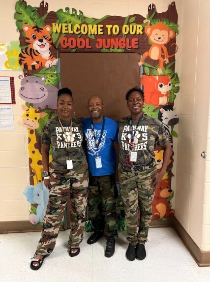 Three people in matching camouflage pants and ID lanyards stand by a decorated door.