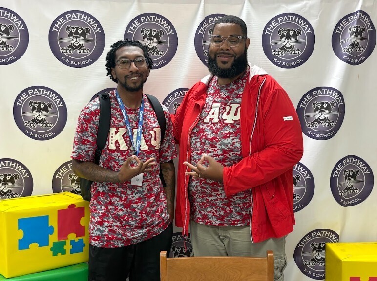 Two men in printed shirts, one with a backpack, pose next to a wall with logos and a yellow puzzle box.