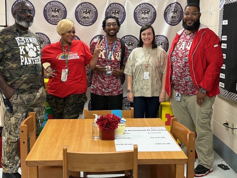 Five people in a classroom. A table with a red flower centerpiece and a paper is in the foreground.