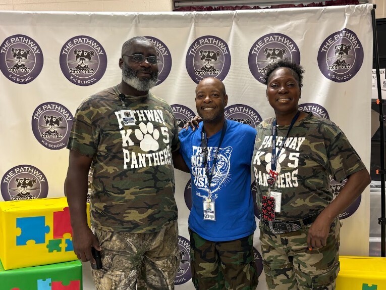 Three people in camouflage uniforms stand near a white banner with logos and text. A large multicolored box is on the left.