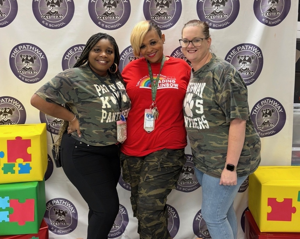 Three women in matching camouflage shirts stand before a backdrop with logos and colorful puzzle pieces.