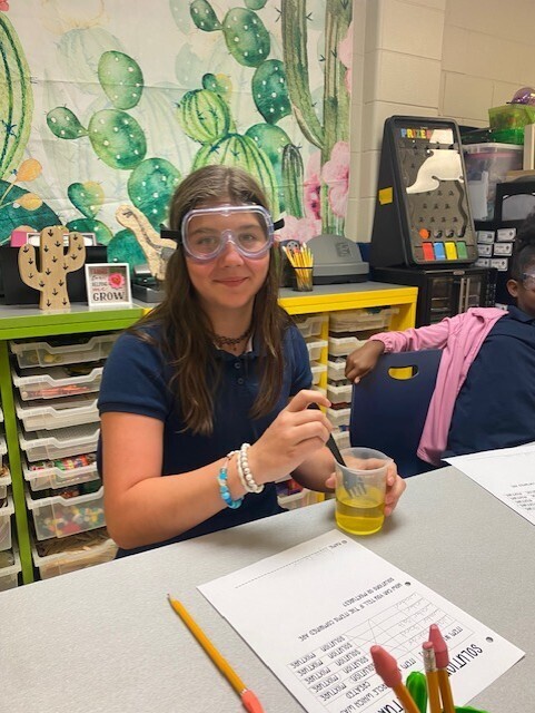 A girl in goggles mixes a yellow liquid in a cup, seated at a desk with pencils, papers, and a cactus mural in the background.
