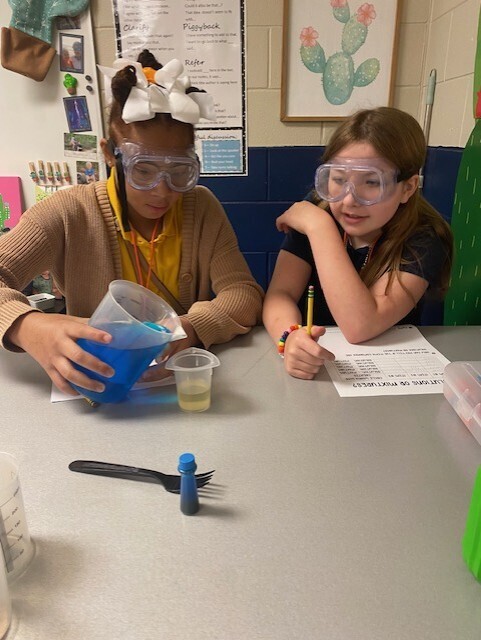 Two students wearing goggles conduct a science experiment. One holds a blue liquid, and the other writes on paper.