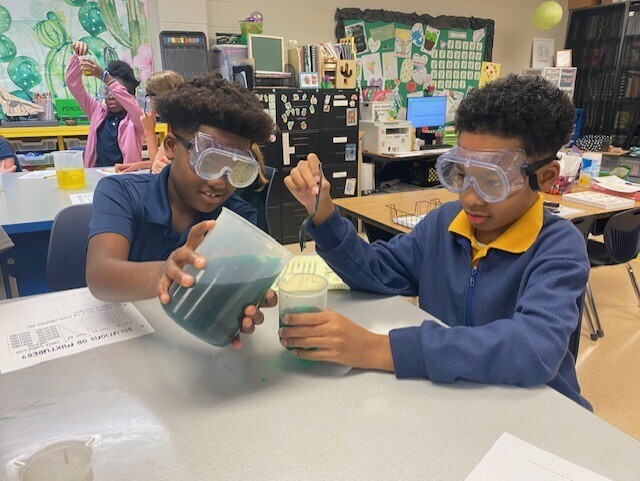 Two children in goggles mix liquids in beakers at a table in a classroom. Behind them, a teacher raises their hand.