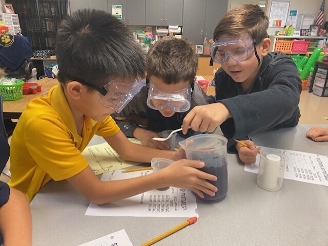 Three students in goggles, seated at a table, with one mixing a substance into a cup.