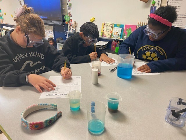 Four students in a classroom are engaged in an activity at a table with beakers and safety goggles.