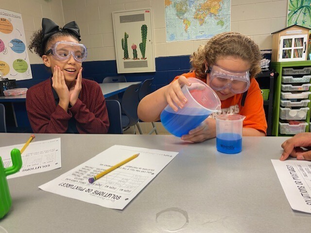 Two students wearing safety goggles perform a science experiment, pouring blue liquid into a beaker.