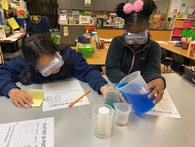Two students wearing safety glasses and masks are working on a chemistry experiment at a table.