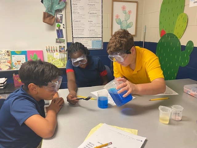 Three children at a table with goggles on. One child pours blue liquid from a beaker into a cup.