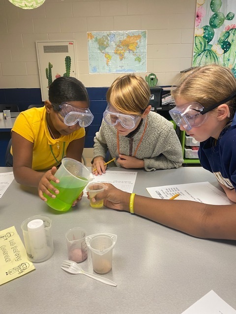 Three children in goggles mix chemicals in a classroom. Papers and various lab materials are on the table.