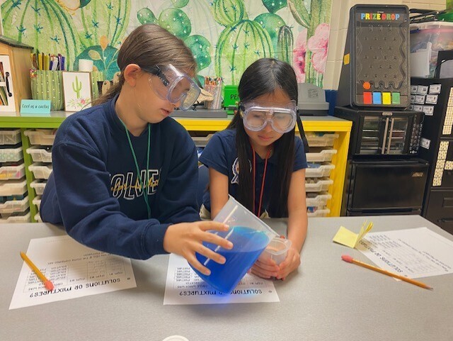 Two students wearing goggles work on a science experiment with blue liquid in a plastic bag.