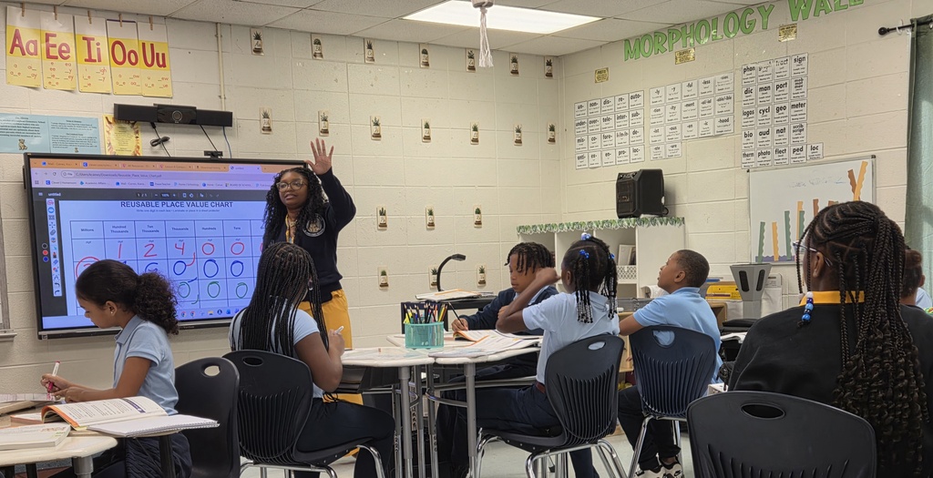 A woman speaks in front of a screen. Children sit in a classroom with chairs and tables.