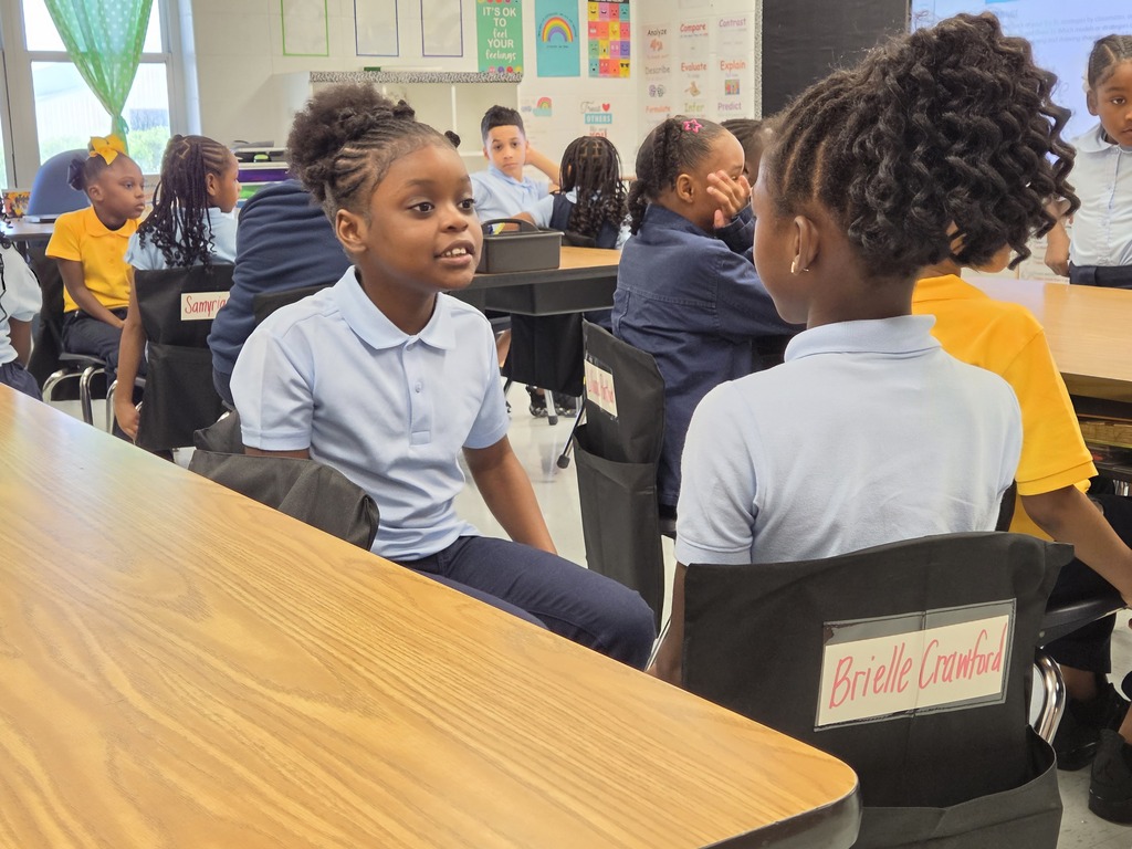 Children sit in chairs in a classroom, with one girl looking upward. A desk and chairs surround them, and others sit in the background.