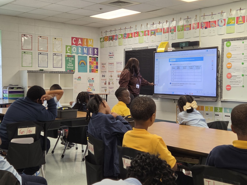 A classroom with children seated around desks. A teacher stands in front of a large screen showing a presentation.