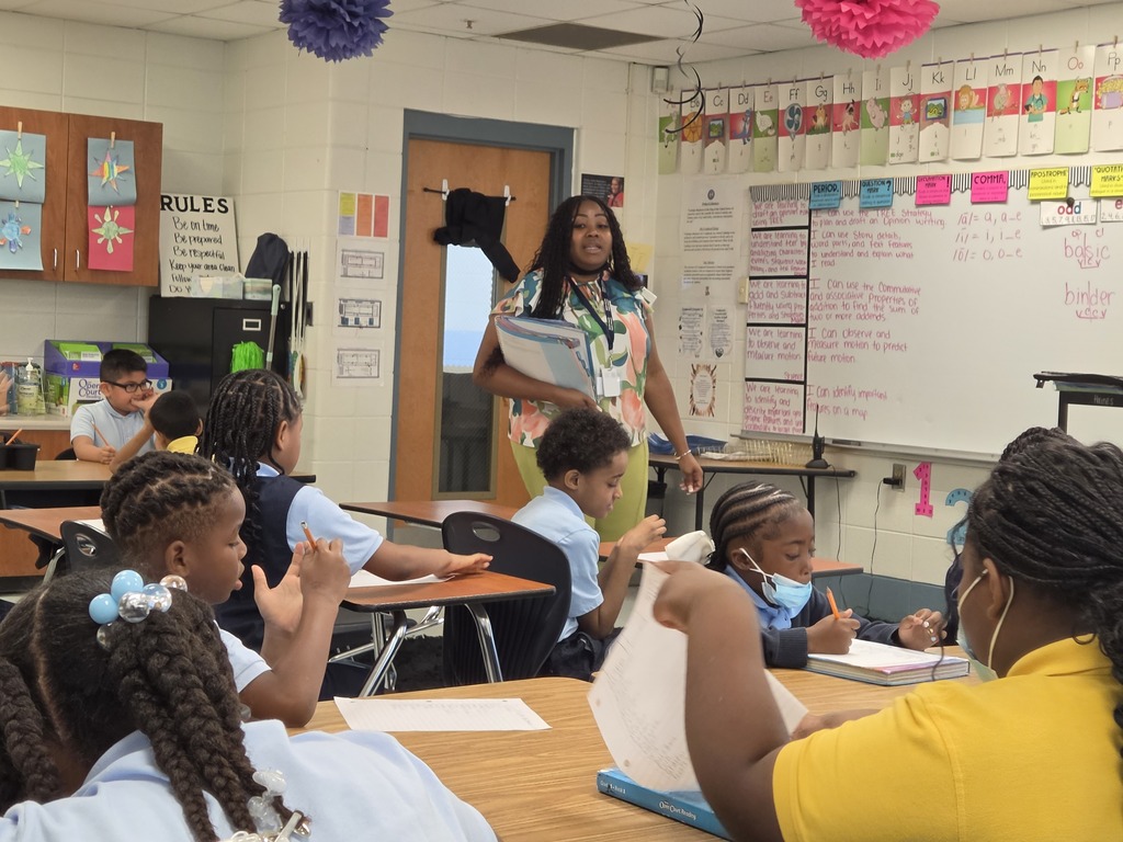 A classroom with students seated. A teacher stands before them, holding a folder. A whiteboard displays colorful papers and notes.