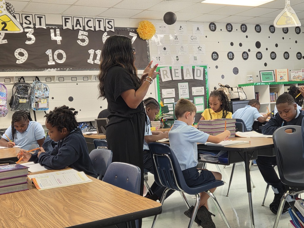 Classroom with teacher standing by wall with "Fast Facts" banner. Students seated at desks, engaged in work. Desks, chairs, backpacks.