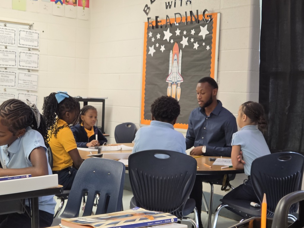 In a classroom, a man reads from a book to a small group of children sitting at a table.