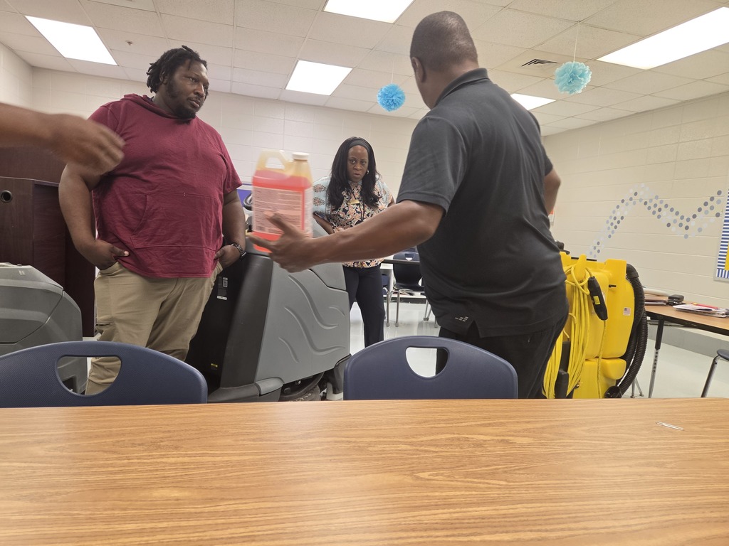 Three people in a room, one holding a bottle. A woman is near a table with chairs. A backpack and a yellow bag are on the floor.