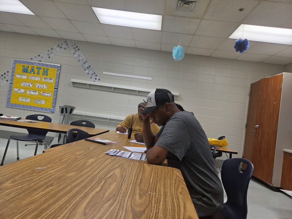 A classroom with tables and chairs. A man sits at a table with his head down, another person behind him. A yellow sign reads "MATH" on the wall.