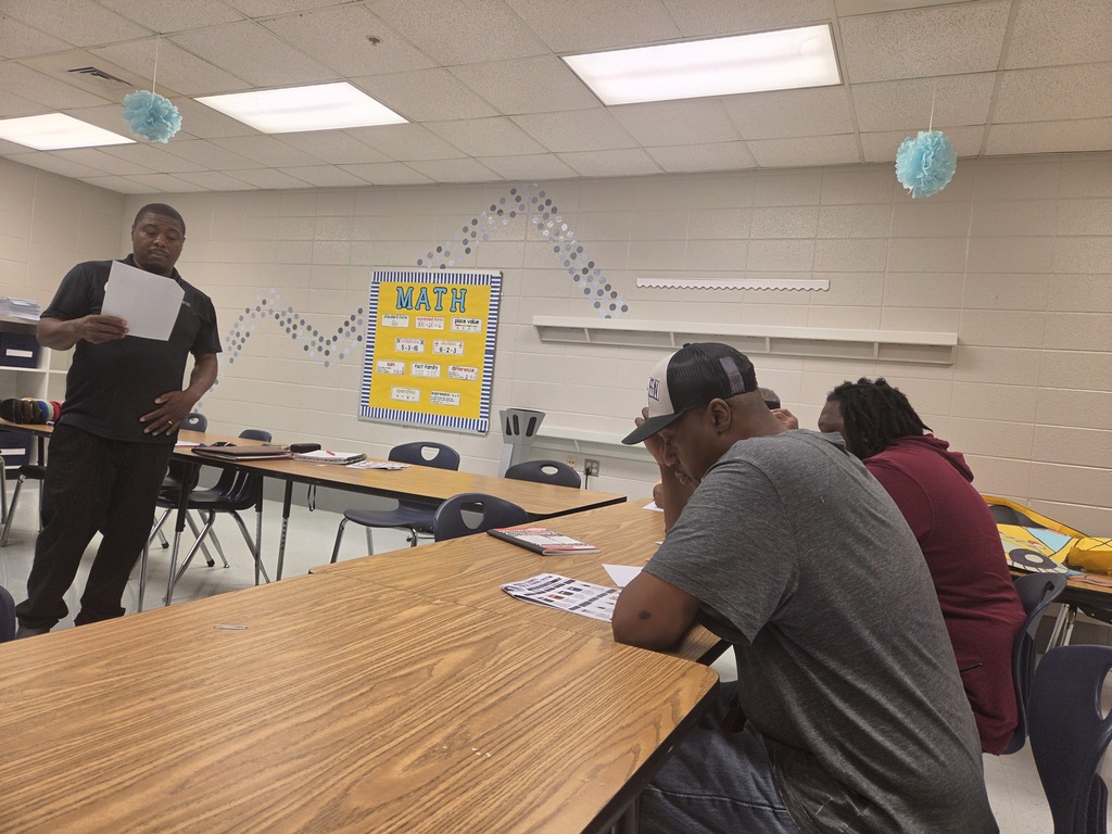 A man stands in a classroom, holding a paper, while a seated man listens. Behind them, a white board displays "MATH".