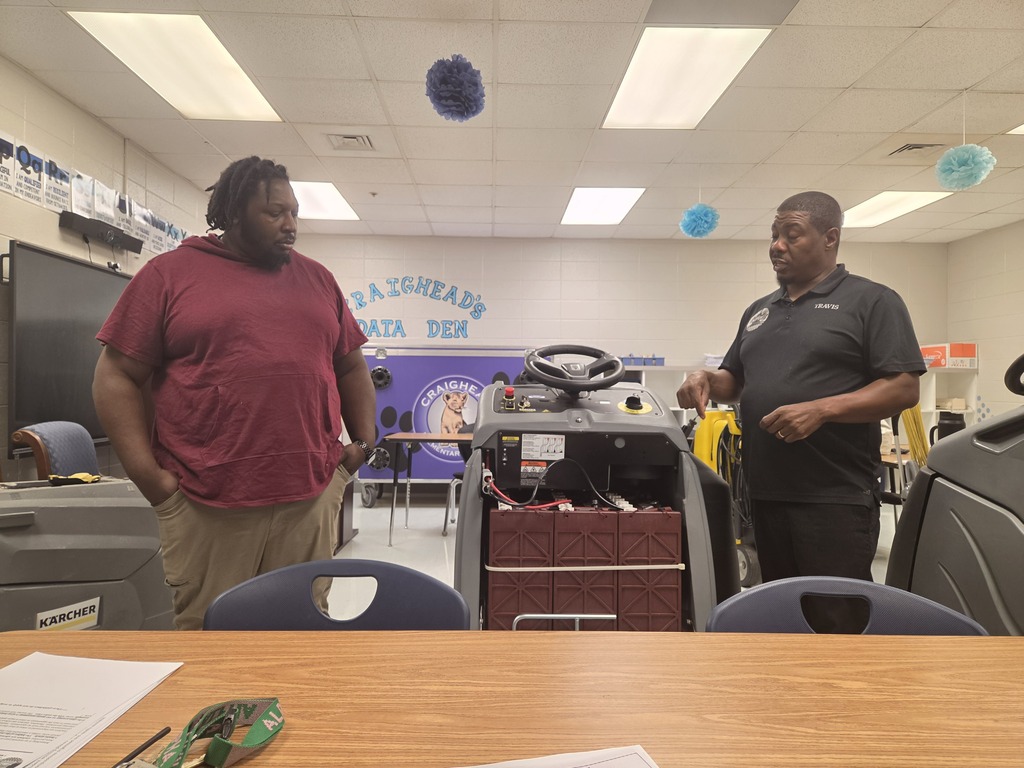 Two men stand in an office, one in a maroon shirt and the other in black, discussing a red and black machine.