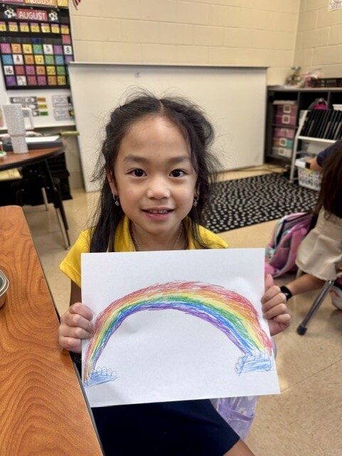 A young girl smiles while holding a white paper with a rainbow drawing in a classroom.