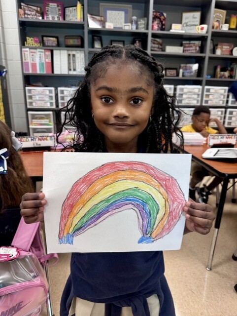 A young girl in a blue dress displays a colorful rainbow drawing. Behind her, a classroom with shelves of books and children at tables.