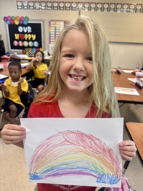Girl holding a colorful drawing of a rainbow in a classroom with other children.