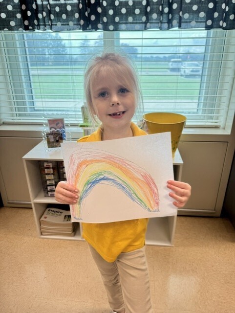 A young girl in a yellow shirt holds a colorful rainbow drawing, standing in a room with a white shelf and window.