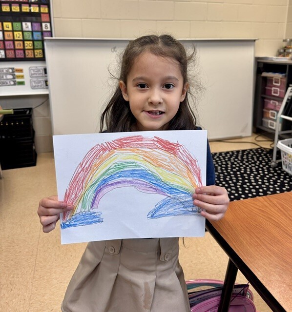 A young girl with a brown dress displays a colorful drawing of a rainbow. The scene is indoors.