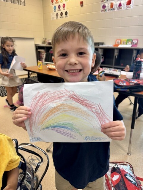A young boy displays a drawing of a colorful rainbow. A classroom with tables and chairs is in the background.