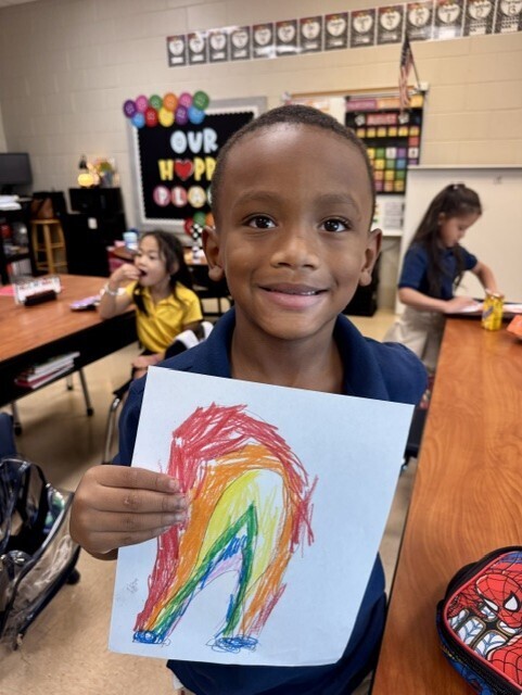 A smiling young boy in a blue shirt shows a colorful rainbow drawing in a classroom.