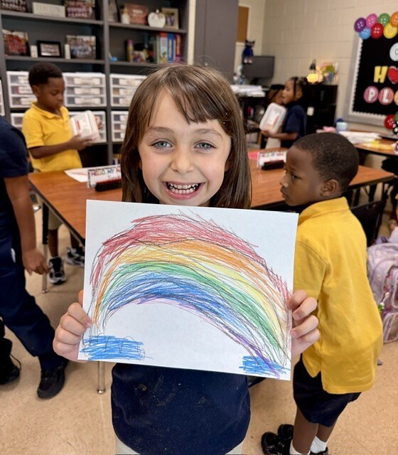 Child holds up rainbow drawing, smiling in classroom, with other children reading and sitting at tables.