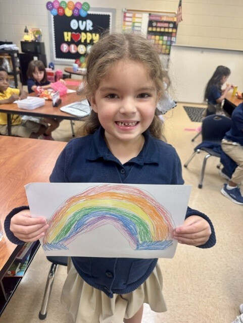 Young child holding a colorful rainbow drawing in a classroom with desks and other students.