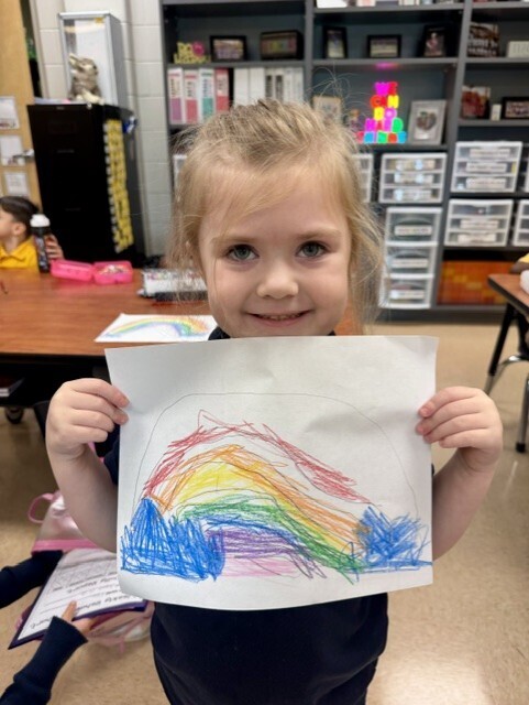 Child holding up a drawing of a rainbow. Behind her, a classroom with tables and shelves.