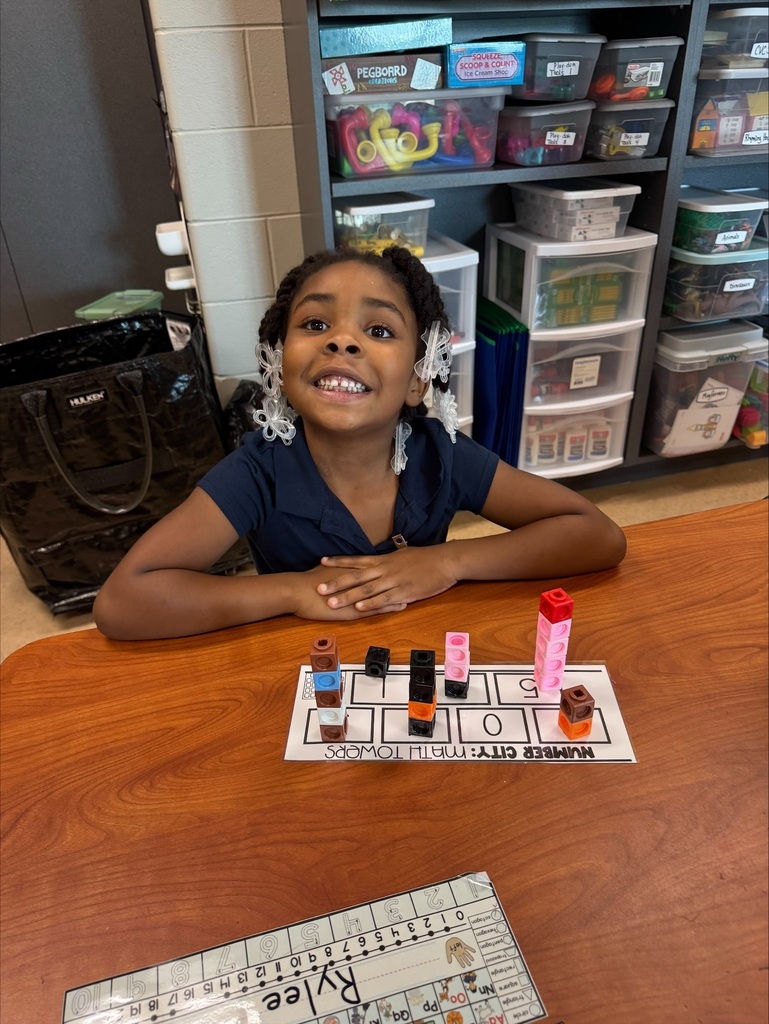 A young girl sits at a table, smiling, with a colorful block arrangement and a number grid nearby.