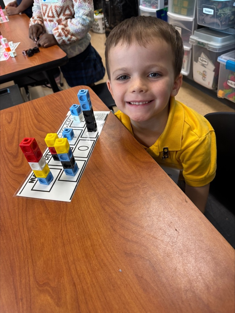 A young boy in a yellow shirt is seated at a table. He is smiling and leaning over a game board with colored pieces.