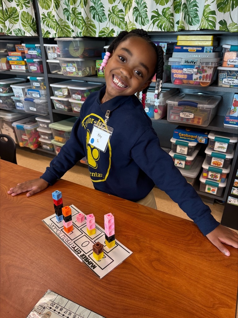 A child smiles while standing behind a table with building blocks and a worksheet. Shelves with storage boxes are behind the child.