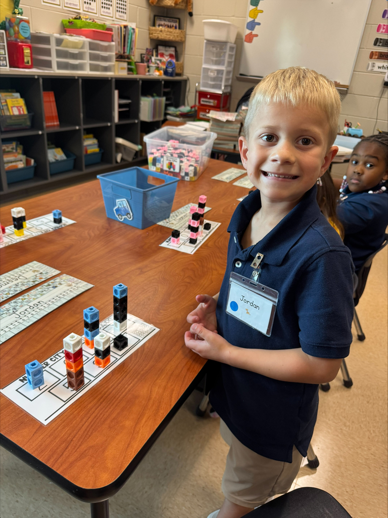 Boy in navy shirt smiles, standing by table with blocks. Behind him, a girl sits at a desk.